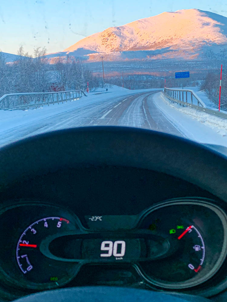 Fahren auf Eisstraßen mit 90 km/h in Nord-Schweden. Die Temperaturanzeige zeigt -23 Grad Celsius an. Aus der Front-Scheibe ist ein verschneiter Hügel zu sehen.