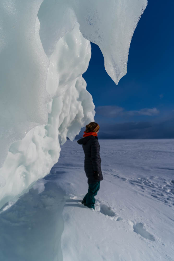 Melanie im Abisko Nationalpark im hohen Norden von Schweden. Sie steht direkt neben einer Eisformation im Schnee und blickt in die Ferne. Der Himmel ist kräftig blau. Sehenswerte Reiseziele in Schweden