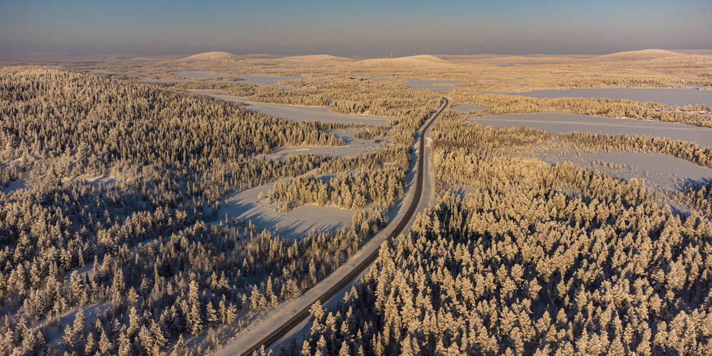 Sehenswerte Reiseziele - Nord Schweden aus Vogelperspektive. Verschneite Waldlandschaft, durch den Wald führt eine Straße. Am Horizont sind ein paar kleinere Hügel zu sehen.