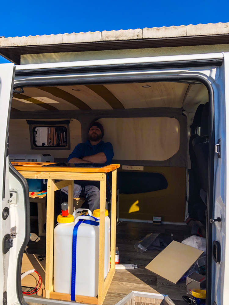 Julian sitzt im Camper und blickt auf die neu ausgebaute Küche im Van. Die Küchenplatte inklusive Induktionskochfeld sitzt, es fehlen noch die Außenwände rund um die Wasserkanister.