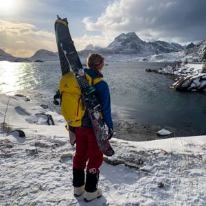 Start Splitboard Tour im Winter auf den Lofoten. Melanie steht am Atlantik im Schnee und blickt auf die Bergkulisse des Fjords. Die Splitboard Bretter sind am Rucksack befestigt.