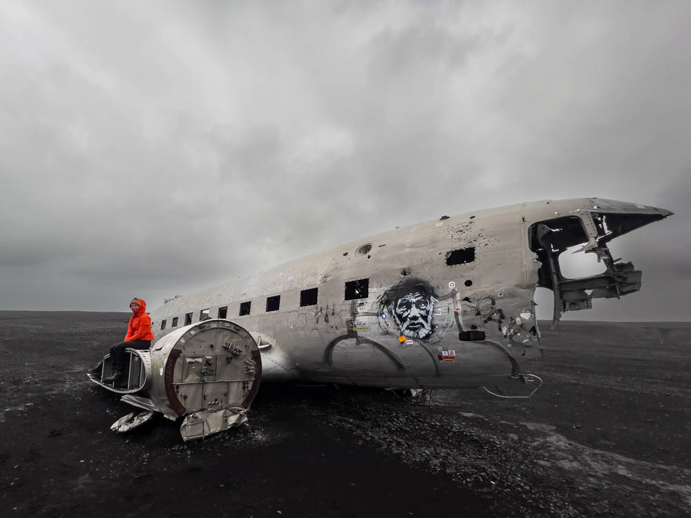 Flugzeugwrack in Island. Melanie sitzt auf dem Triebwerk des Wracks. Düstere Stimmung am Himmel. 5 Jahre Entscheidung zur Weltreise