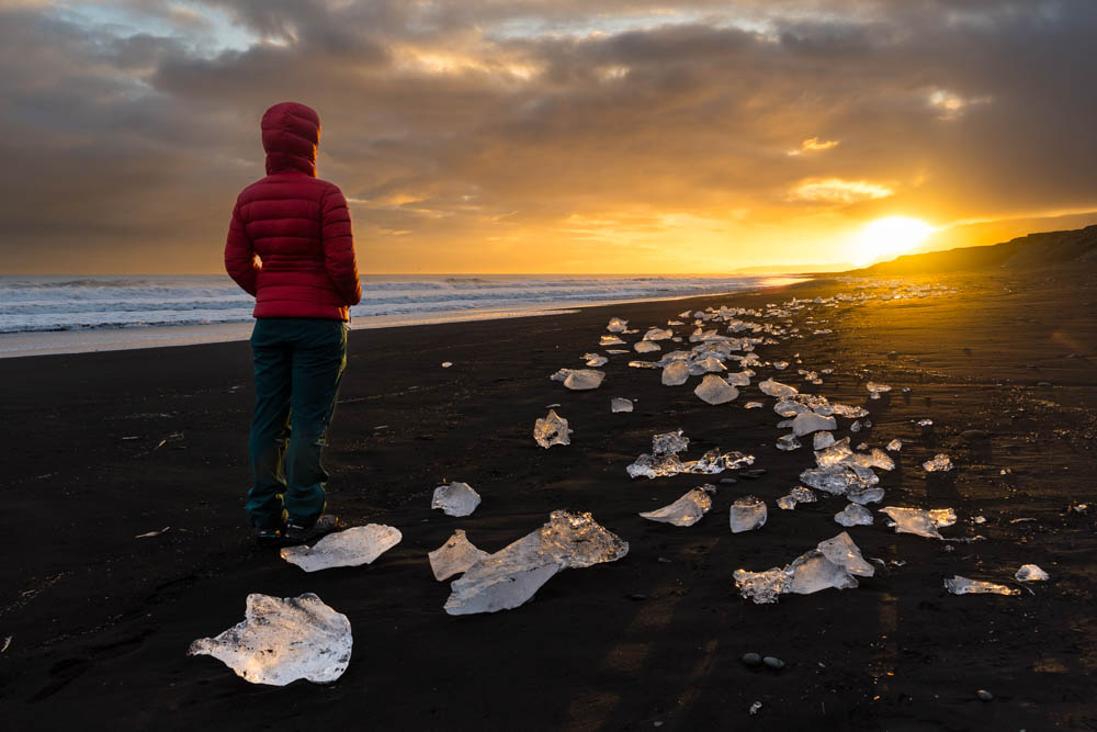 Melanie steht am Diamond Beach in Island im Herbst und beobachtet den Sonnenuntergang. Es liegen kleinere Eisbrocken am Strand, welche von der untergehenden Sonne angestrahlt werden. 5 Jahre Entscheidung zur Weltreise