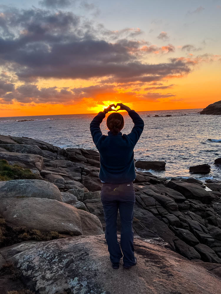 Sonnenaufgang am Meer in Spanien an der Biskaya. Melanie blickt auf das Meer und bildet mit ihren Händen ein Herz vor der orangenen Sonne.