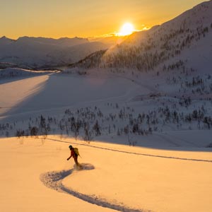 Melanie bei Abfahrt mit Splitboard in verschneiter Berglandschaft. Sonnenuntergang über den Bergen erleuchtet Schnee orangefarben. Tourengehen und Nordlichter auf Senja.