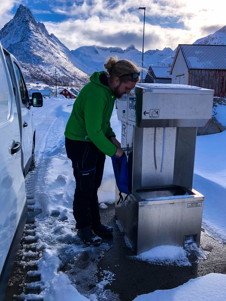 Vanlife auf Insel Senja in Norwegen im Winter. Julian füllt Frischwasser an einer Ver- und Entsorgungsstation. Rund herum liegt Schnee und Van Vivaldi ist zu sehen.