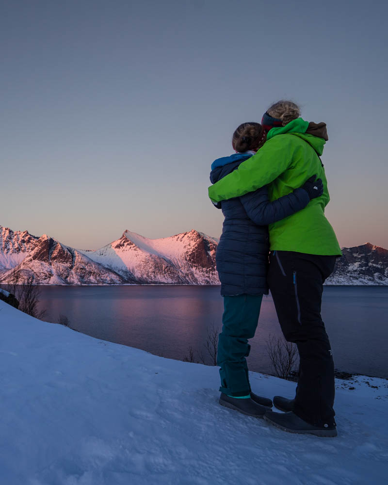 Aufnahme von Melanie und Julian, die sich in den Armen halten und auf den Fjord und die rot leuchtenden Berge schauen. Sonnenuntergang auf Insel Senja in Norwegen