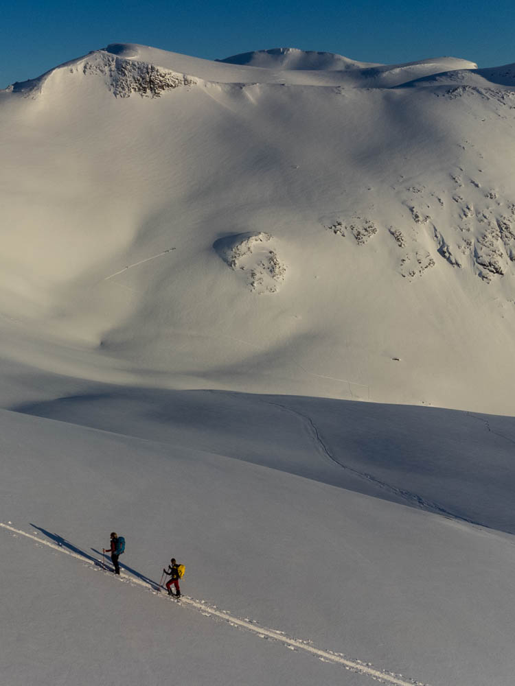 Tourengehen und Nordlichter auf Senja. Drohnenaufnahme von Melanie und Julian im Aufstieg bei Skitour. Blauer Himmel und herrliches Winterwonderland