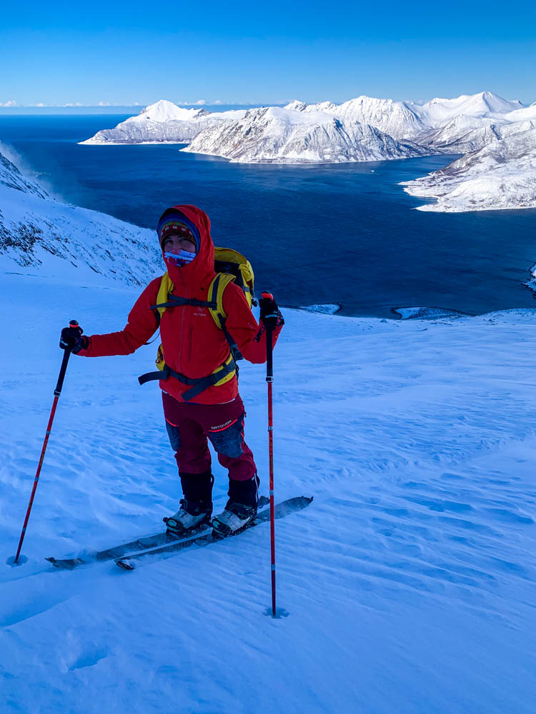 Melanie beim Tourengehen auf Senja. Es ist sehr windig und kalt. Hinter Melanie ist das Meer und ein Fjord zu sehen. Kräftig blauer Himmel