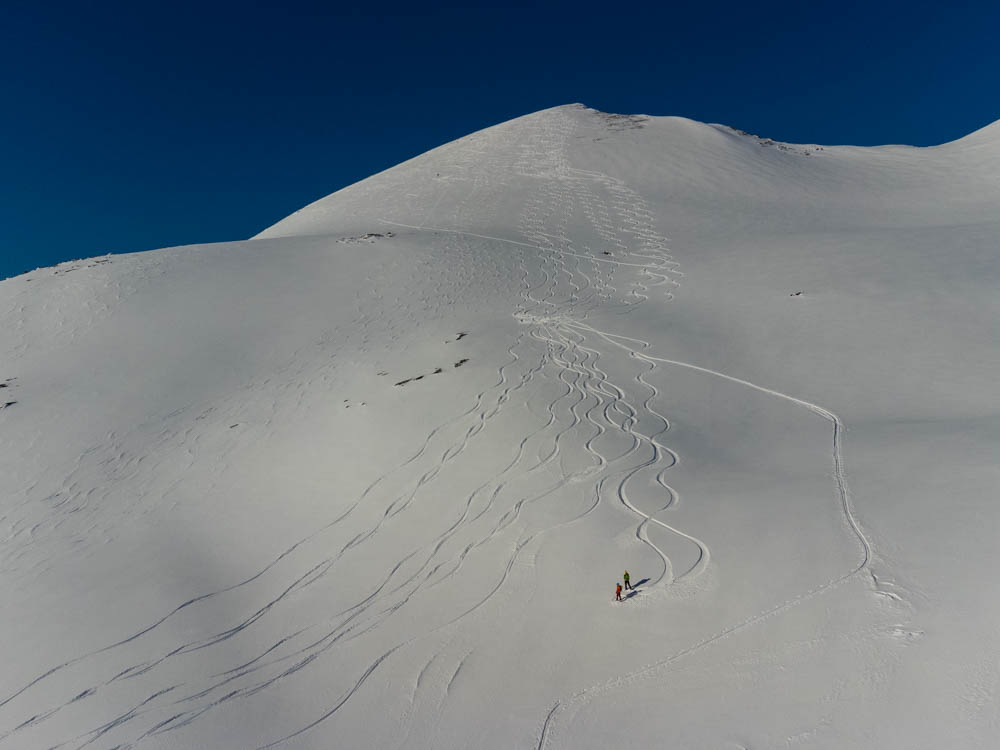 Drohnenaufnahme von Melanie und Julian bei Abfahrt mit Splitboards in den verschneiten Bergen in Senja, Norwegen. Der Himmel ist kräftig blau. Schöne Spuren im Schnee