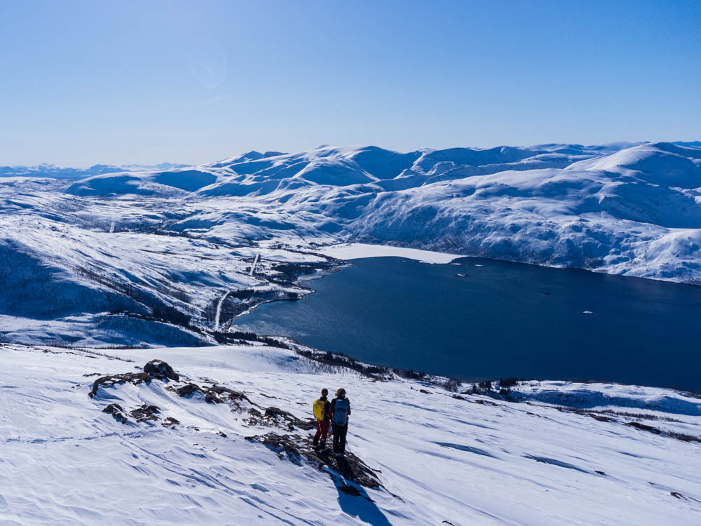 Drohnenaufnahme von Melanie und Julian mit Fjord und verschneiter Berglandschaft im Winter auf Senja, Norwegen. Der Himmel ist kräftig blau.