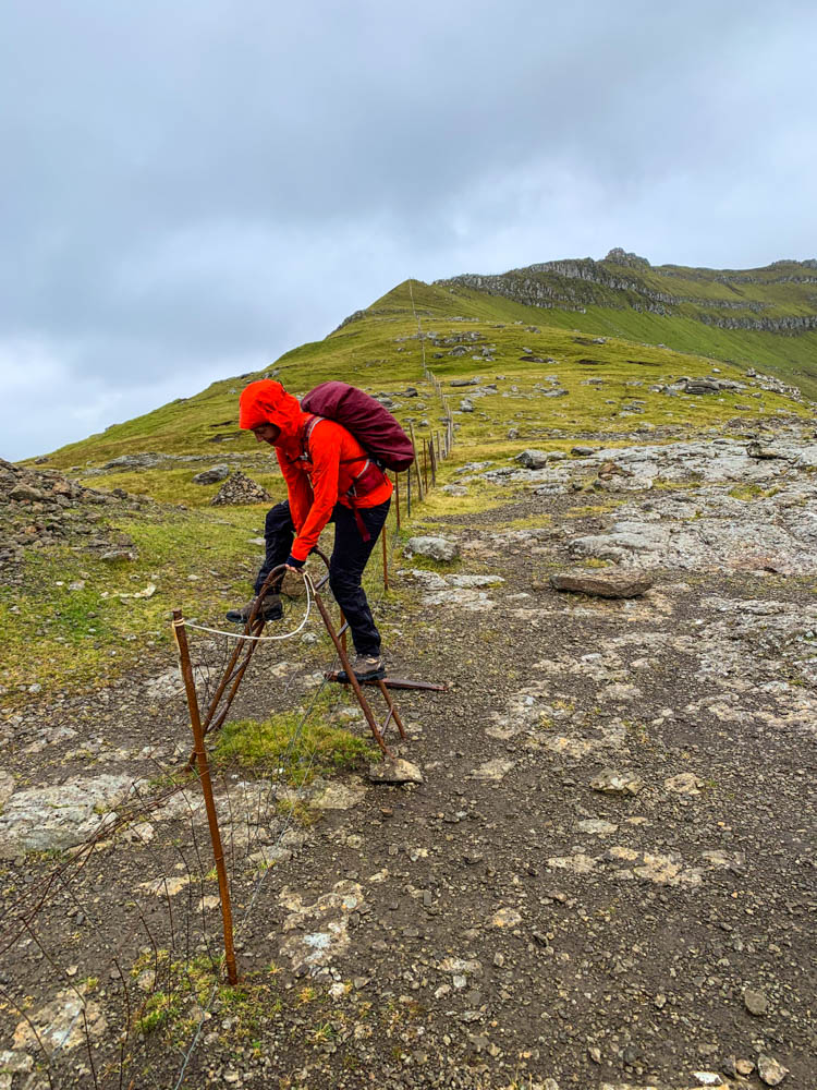 Wanderung von Hellurnar nach Fuglafjørður. Melanie klettert über Zaun mit Trittleiter. Färöer Inseln: Gradlandschaft, Hügel, Regen