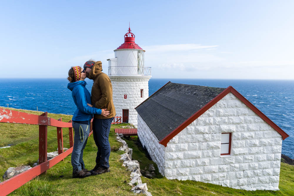 Melanie und Julian küssend vor Leuchtturm Borðan auf Insel Nólsoy der Färöer. Hinter dem wunderschönen Leuchtturm ist direkt das Meer zu sehen sowie der blaue Himmel.