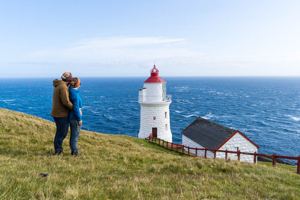 Melanie und Julian mit Leuchtturm Borðan auf Insel Nólsoy. Hinter dem wunderschönen Leuchtturm ist direkt das Meer zu sehen sowie der blaue Himmel.