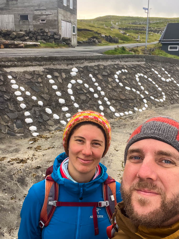 Selfie von Melanie und Julian auf Insel Nólsoy mit Schriftzug "Nólsoy" in der Strandmauer hinter ihnen.