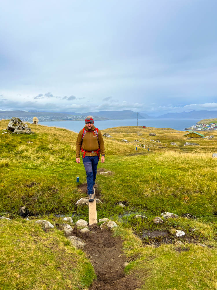 Julian beim wandern auf Insel Nólsoy, Färöer Inseln. Er balanciert über einen schmalen Holzsteg, hinter ihm ist ein Dorf sowie das Meer zu sehen.
