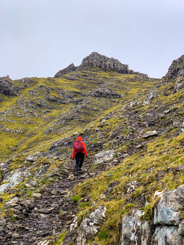 Wanderung auf alter Postroute von Bøur nach Gásadalur auf Insel Vágar der Färöer Inseln. Melanie wandert im steilen Gelände durch Gras- und Felslandschaft bei regnerischen Bedingungen. Mit dem Camper gut zu erreichen.