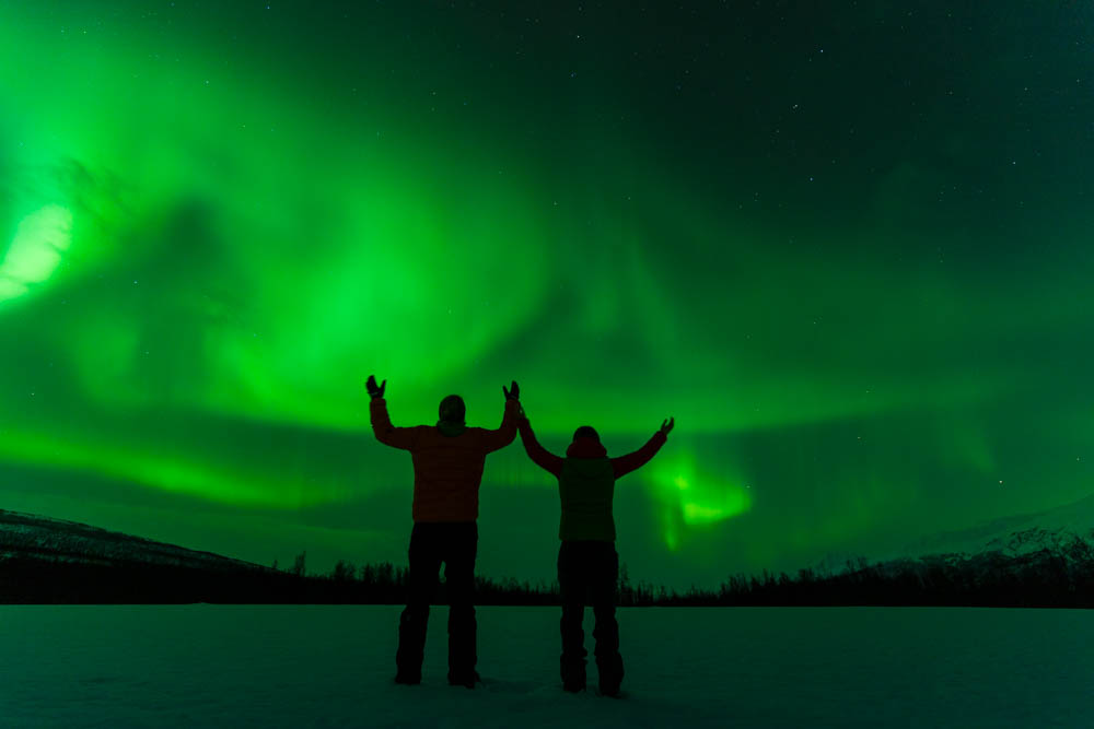 Faszination Nordlichter, Melanie und Julian stehen auf einem Schneefeld und heben die Hände in die Höhe. Über ihnen tanzen die Polarlichter. Winter in Norwegen, Lyngenalpen