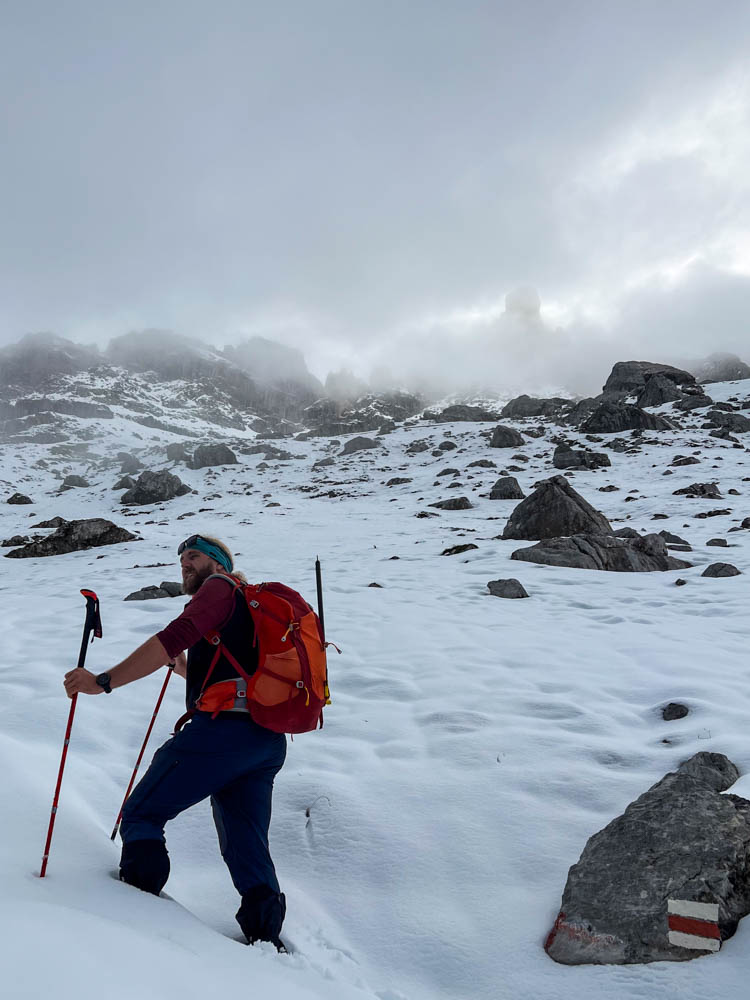 Julian bei einer Wanderung im Schnee. Er hat Stöcke in den Armen und blickt auf die schöne Alpenlandschaft, welche ein wenig in Wolken liegt. Packliste für eine Tageswanderung