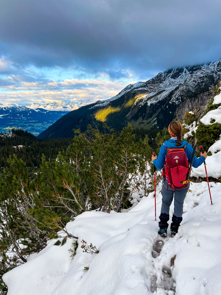Melanie bei einer Wanderung im Schnee. Sie hat Stöcke in den Armen und blickt auf die schöne Alpenlandschaft. Packliste für eine Tageswanderung