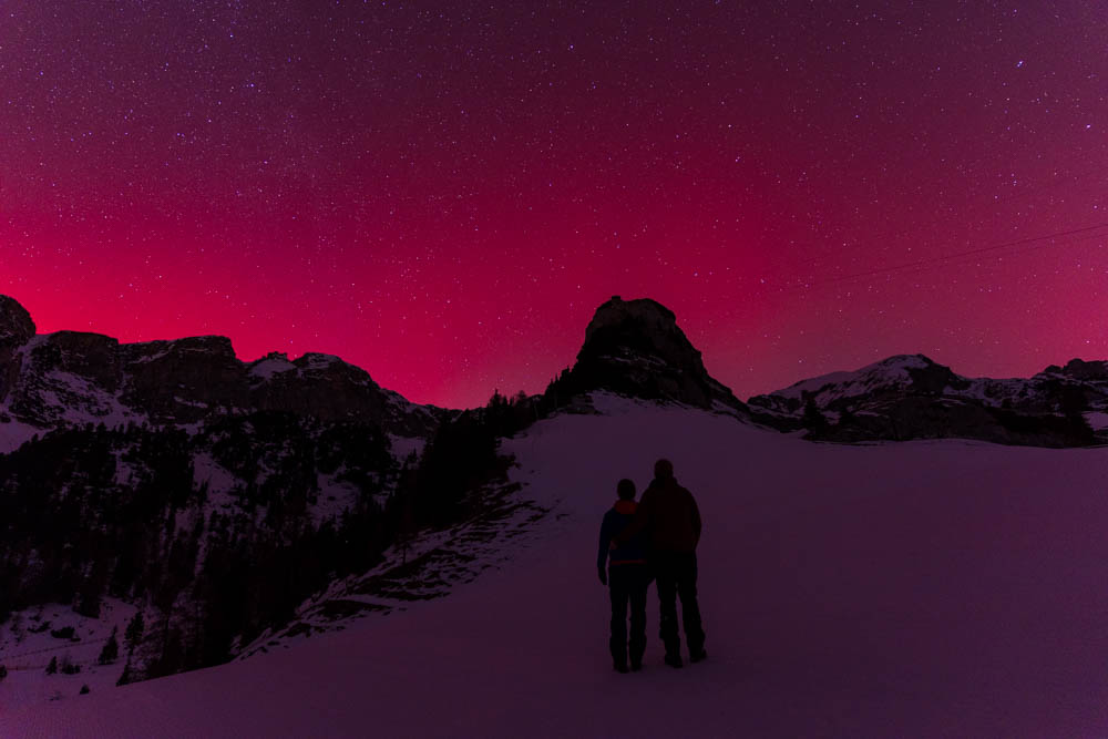 Polarlichter in den Alpen über dem Achensee. Der Himmel leuchtet violett über dem Rofangebirge. Melanie und Julian stehen im Schnee der Skipiste und bestaunen das Naturschauspiel.